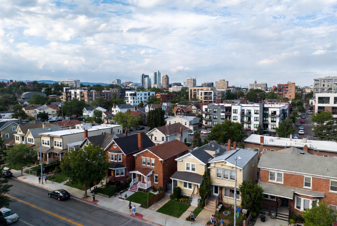 Aerial view of a residential neighborhood with houses and mid-rise apartments, with a city skyline in the distance under a partly cloudy sky.