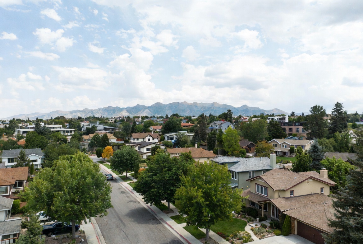 Aerial view of a tree-filled suburban neighborhood with homes and streets, with a mountain range on the horizon under a partly cloudy sky.