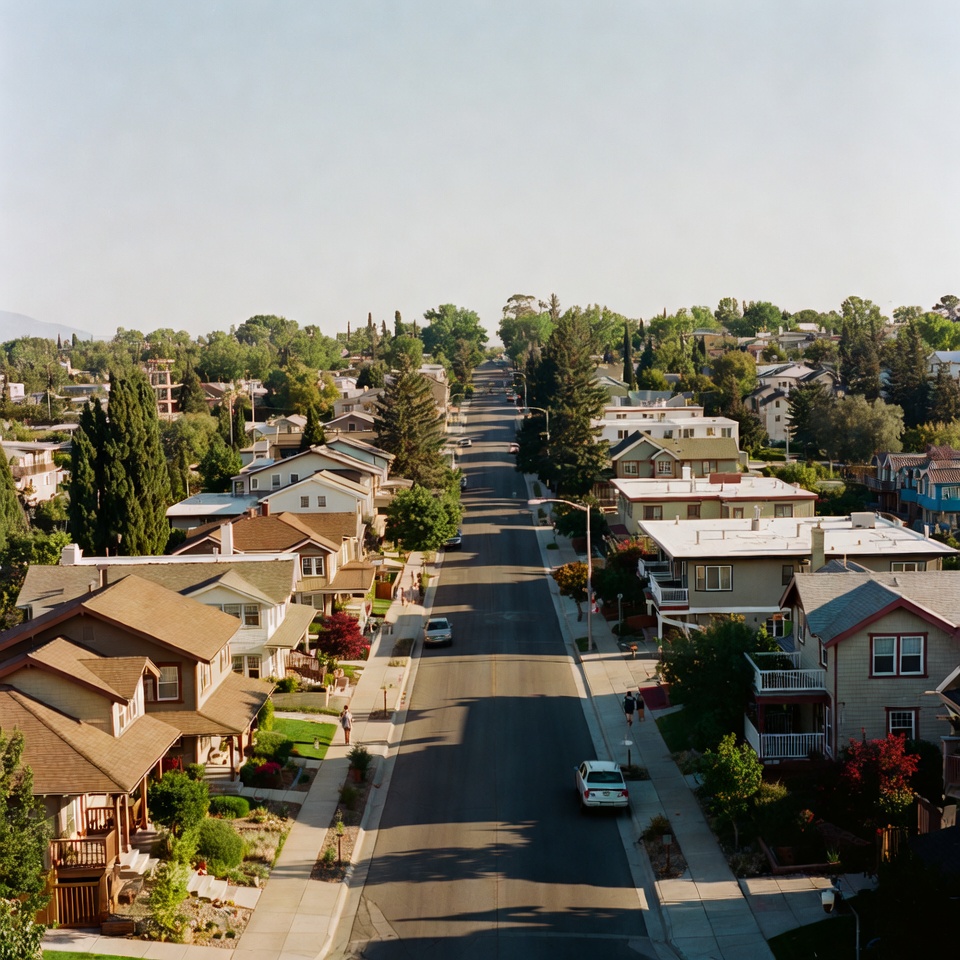 Aerial view down a long residential street lined with homes, trees, and parked cars in warm late-day light.
