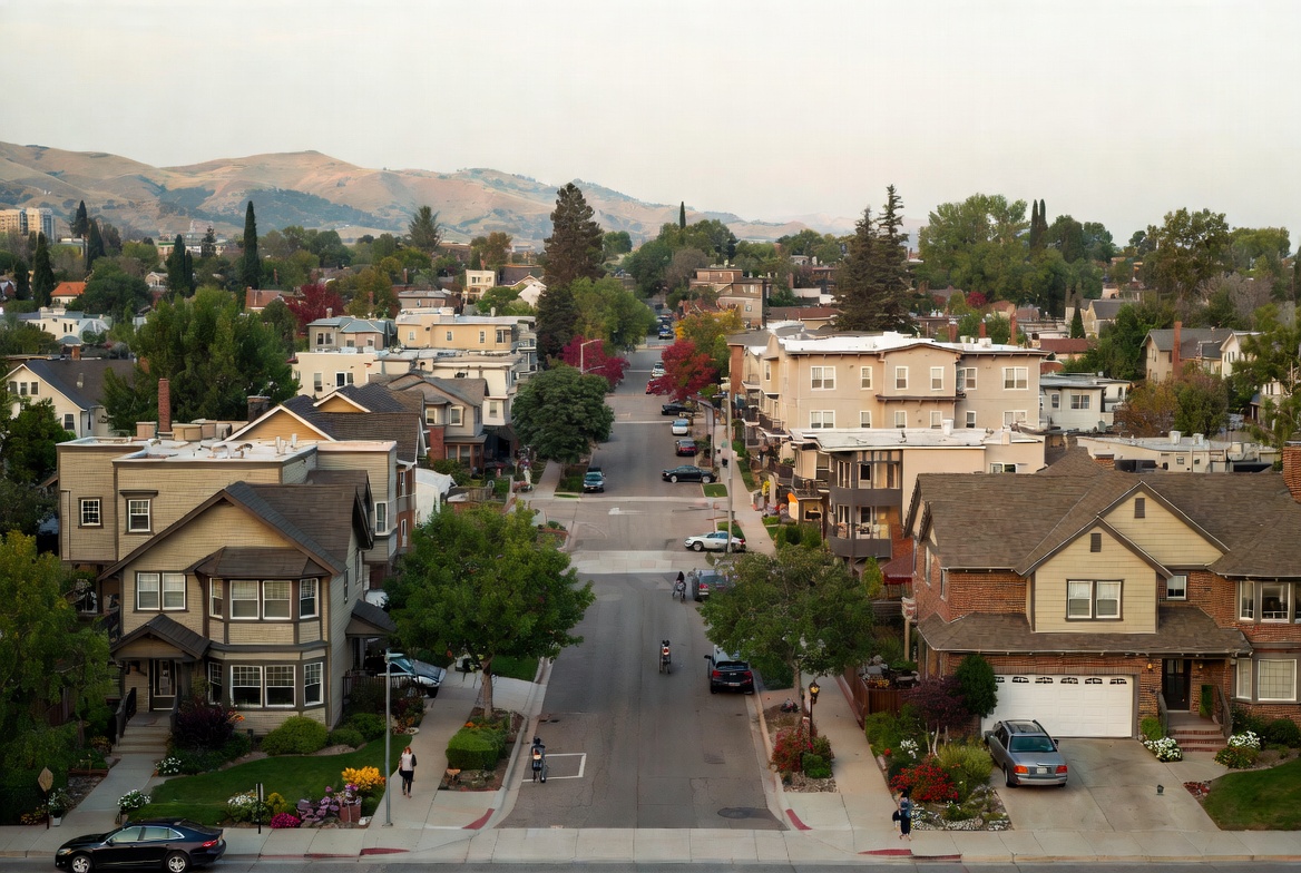 Aerial view down a residential street lined with homes and trees, with rolling hills in the distance under a pale evening sky.