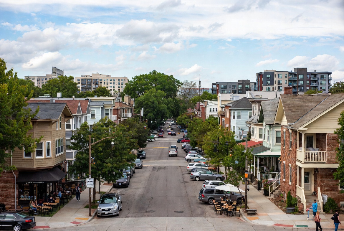 Tree-lined city street with parked cars and sidewalk dining, with mid-rise apartment buildings in the distance under a partly cloudy sky.