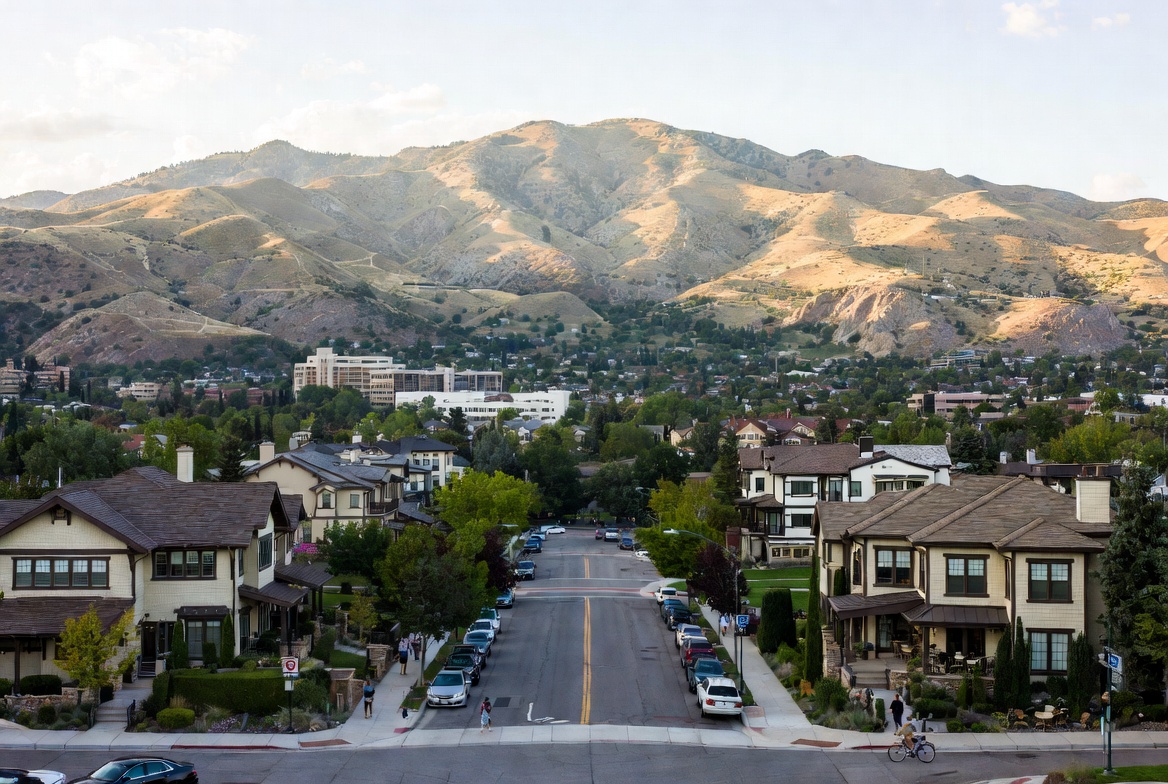 Residential street lined with homes and parked cars, leading toward sunlit mountains in the background under a pale sky.