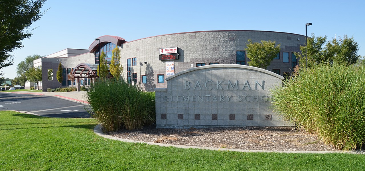 Single storey school building with a curved metal roof and tan brick walls, with a stone monument sign in the foreground reading Backman Elementary School, set among lawn and ornamental grasses.
