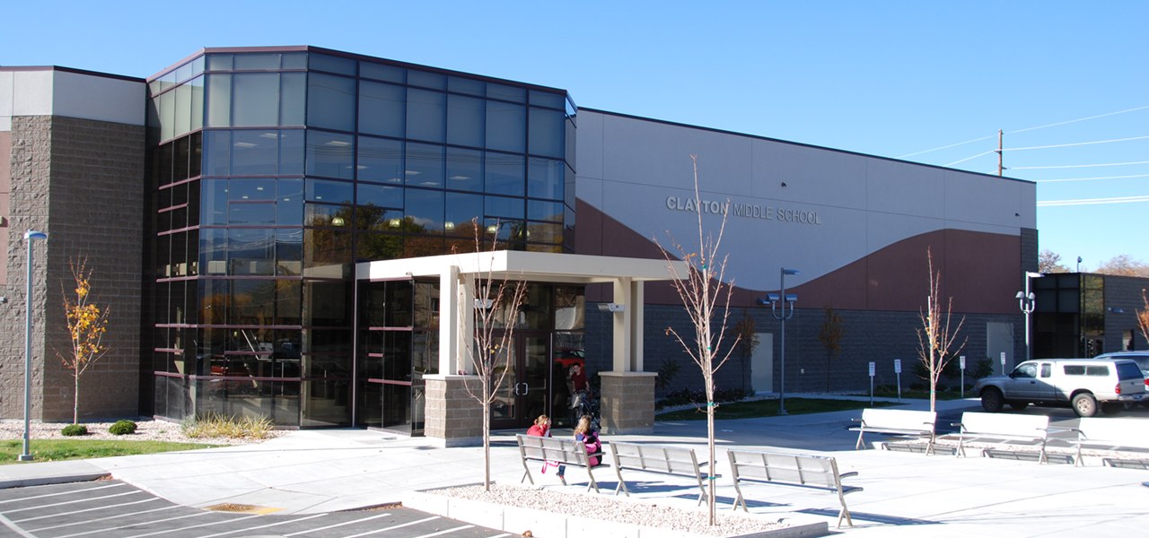 Modern school building with a two storey angled glass curtain wall, a white flat entrance canopy and a maroon painted wave panel carrying the words Clayton Middle School, with benches and bare trees on a concrete forecourt.