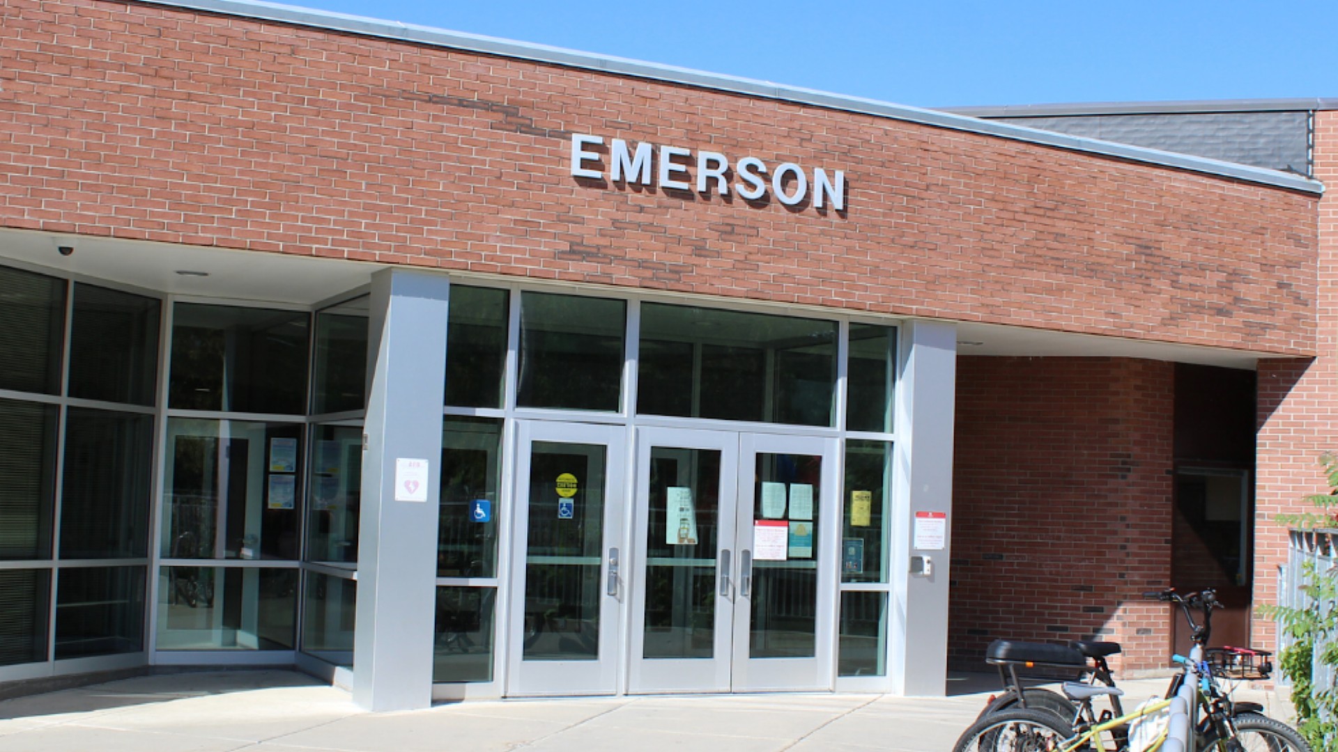 Red brick school building with the word Emerson in raised metal letters above a glass double door entrance, with bicycles parked at a rack on the right under a clear blue sky.