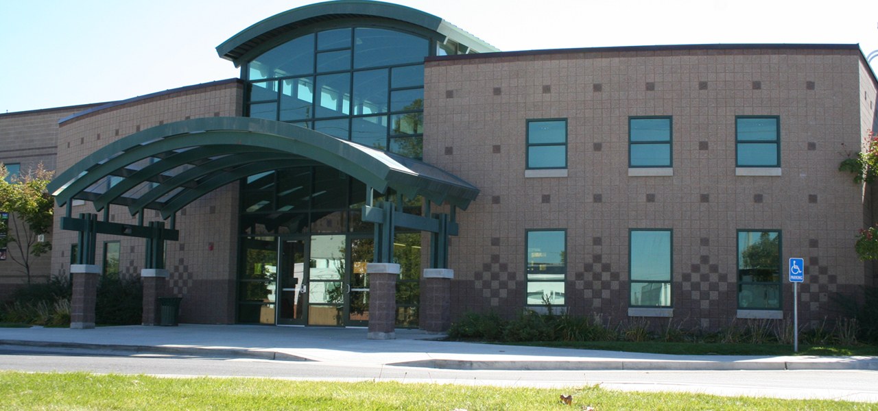 Two storey school building faced in tan and maroon checkered block, with a curved green steel canopy on posts leading to glass entrance doors and a tall glazed stair bay above.