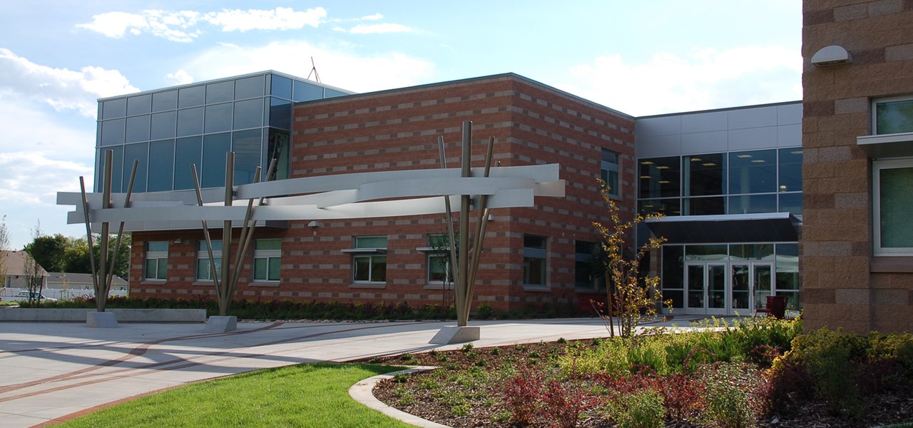 Modern two storey school with red brick and blue glass walls, a horizontal steel trellis on angled posts across the front, and a landscaped bed of shrubs beside a wide concrete plaza.