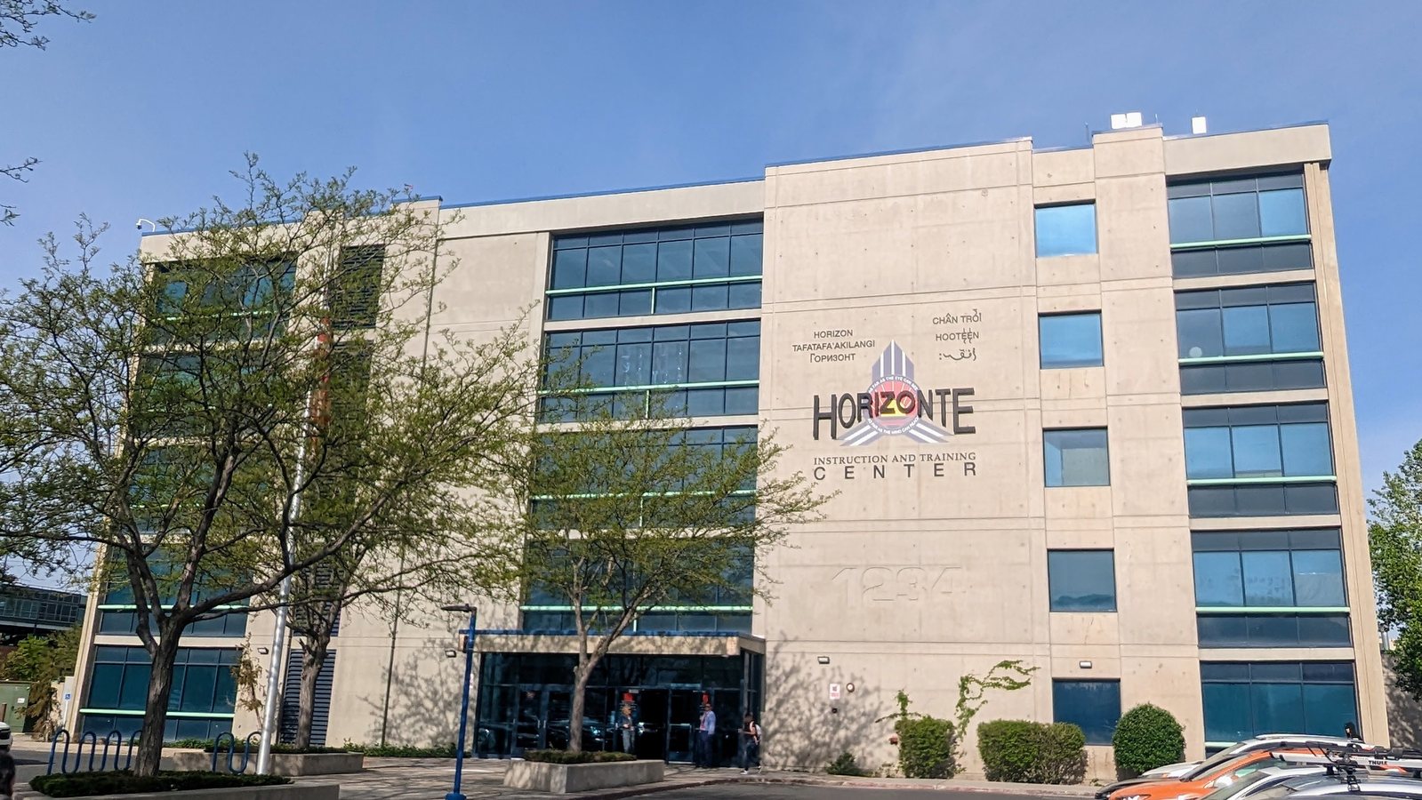 Five storey pale concrete building with dark ribbon windows and large wall lettering reading Horizonte Instruction and Training Center, with the same name repeated in several other languages above it, seen past bare spring trees.