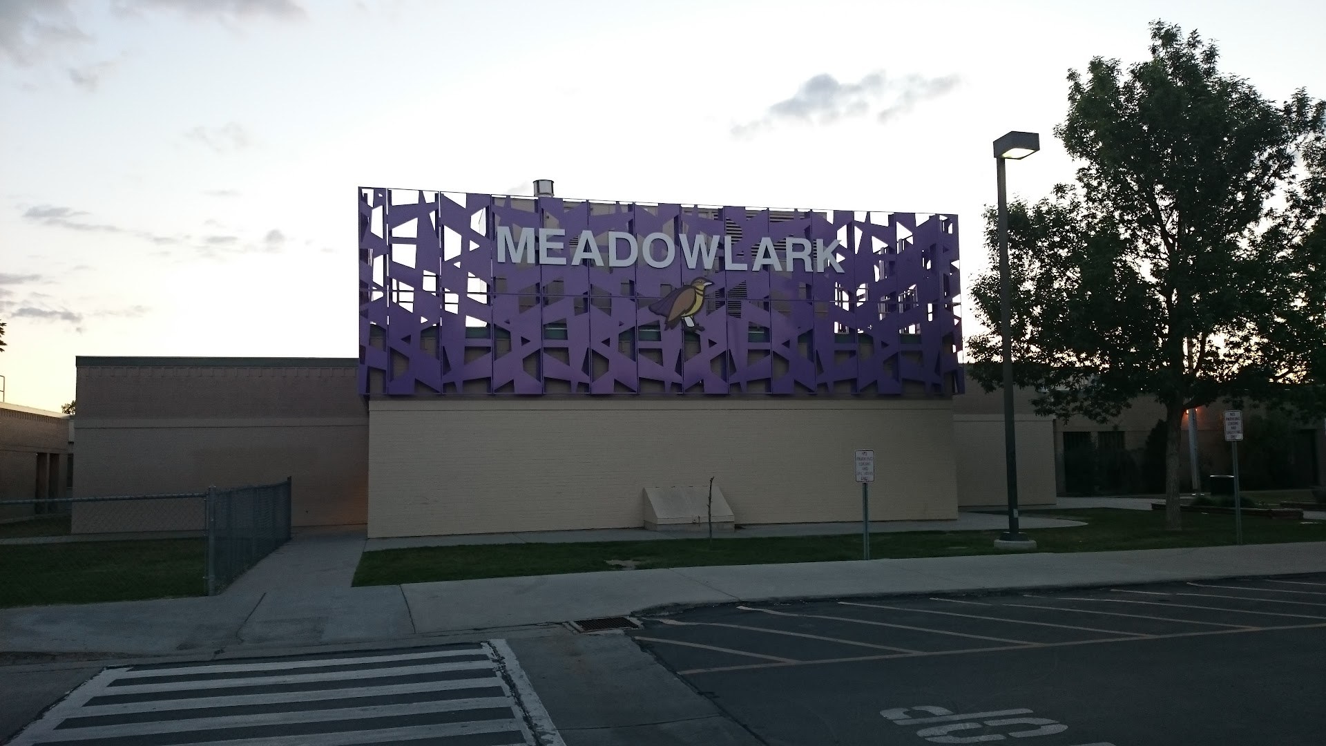 Low tan block school wall topped by a tall purple perforated metal screen carrying the word Meadowlark in white letters beside a yellow and brown bird emblem, photographed from the street at dusk.