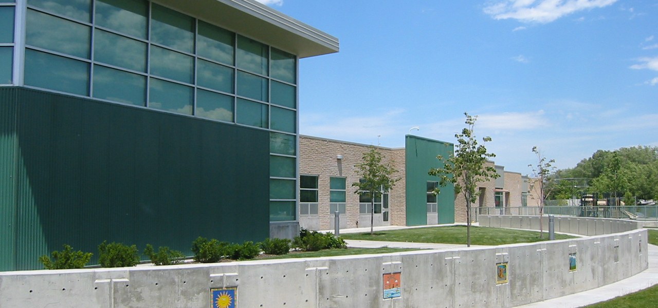 School building with a tall dark green metal clad wing and banks of square windows beside pale block classrooms, fronted by a low concrete retaining wall set with small painted tile artworks.