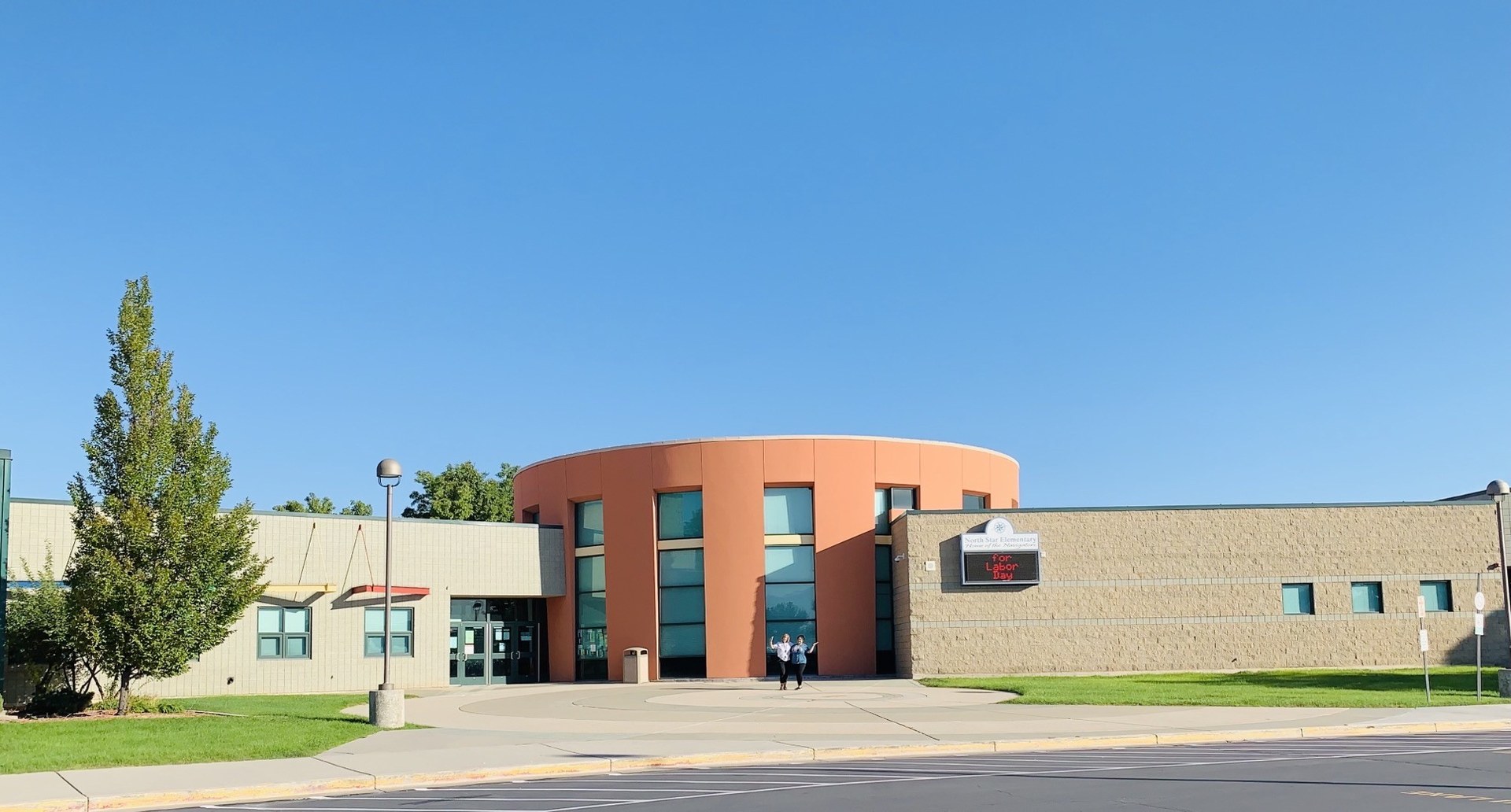 School with a tall orange cylindrical entrance rotunda between low tan block wings, a wall mounted sign to the right reading North Star Elementary Home of the Navigators, and an empty car park in front.