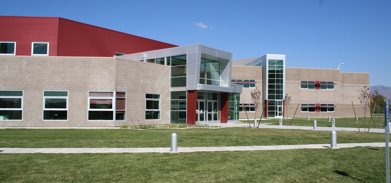 Modern two storey school building with tan block walls, a red panelled upper section and a glass entrance tower, fronted by a mown lawn and young trees.