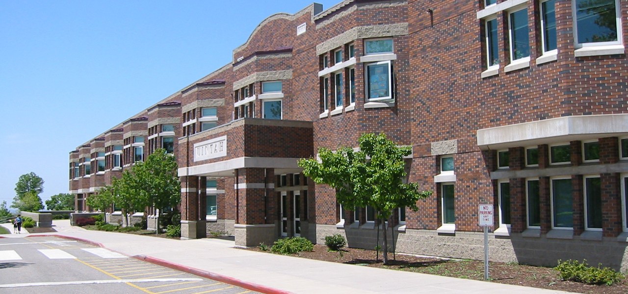 Long two storey brick school building with cast stone banding and a projecting entrance porch carrying inset letters reading Uintah, seen down a sloping pavement beside a kerbside car park.