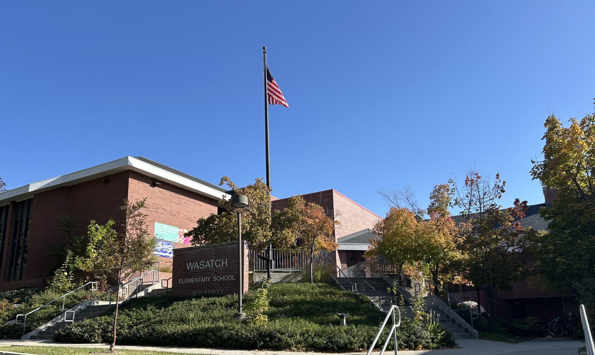 Red brick school on a raised terrace behind autumn trees, with an American flag on a tall pole and a pale monument sign in the foreground reading Wasatch Elementary School 30 R Street.