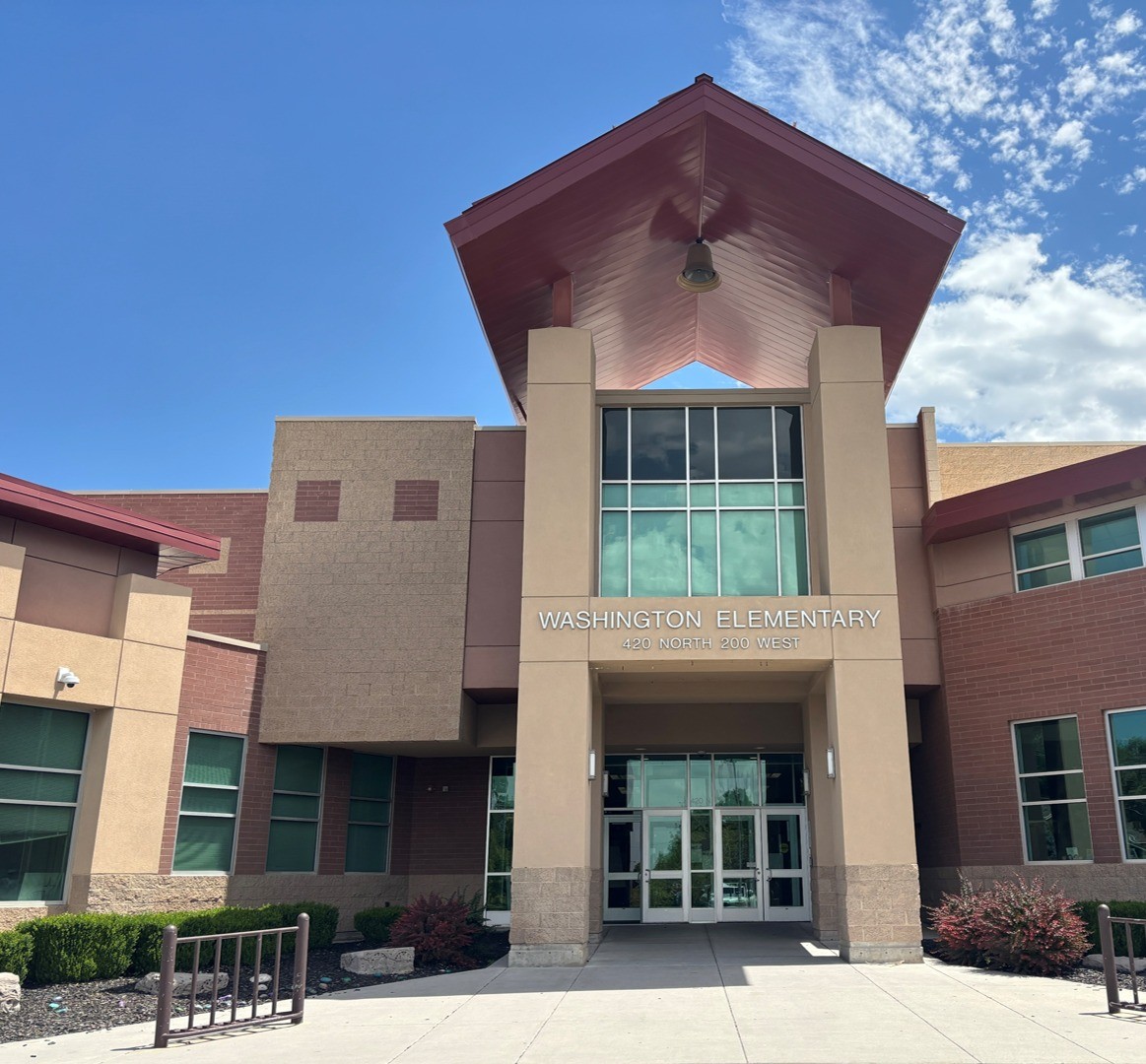 Tall stucco and brick school entrance with a peaked red roof canopy and a hanging lamp, with lettering reading Washington Elementary and 420 North 200 West above the glass doors, and clipped shrubs flanking a concrete walkway.