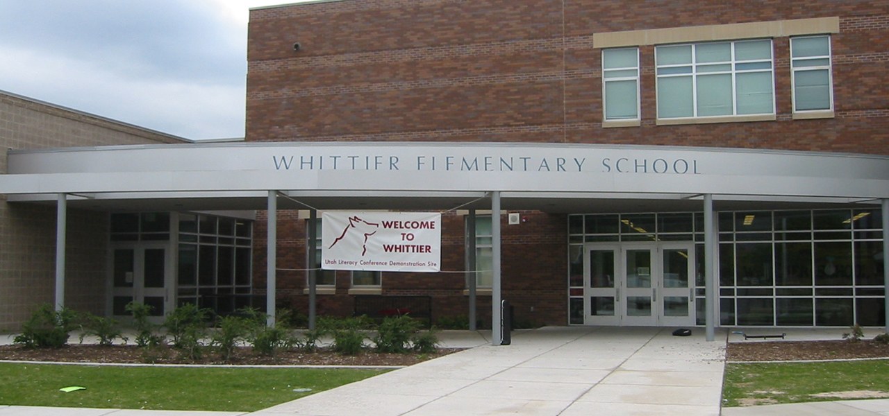 Brick school entrance under a long curved metal canopy carrying the words Whittier Elementary School, with a fabric banner beneath it reading Welcome to Whittier beside a wolf emblem.